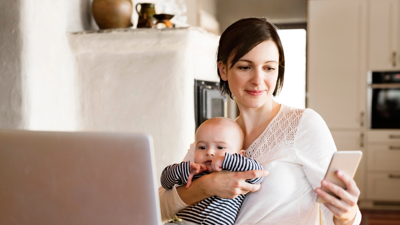 Eine junge Frau sitzt am Küchentisch und telefoniert. Vor ihr steht eine Tasse und ein geschlossenes Notebook.