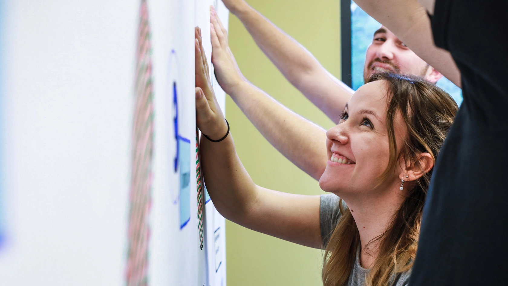Junge Frau steht während eines Seminars mit weiteren Personen an einer Metaplanwand und schaut sich lächelnd die Aufschrift an.