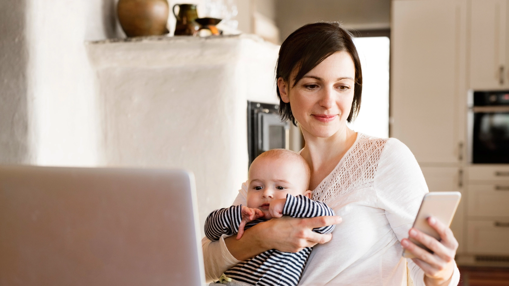 Eine junge Frau sitzt am Küchentisch und telefoniert. Vor ihr steht eine Tasse und ein geschlossenes Notebook.