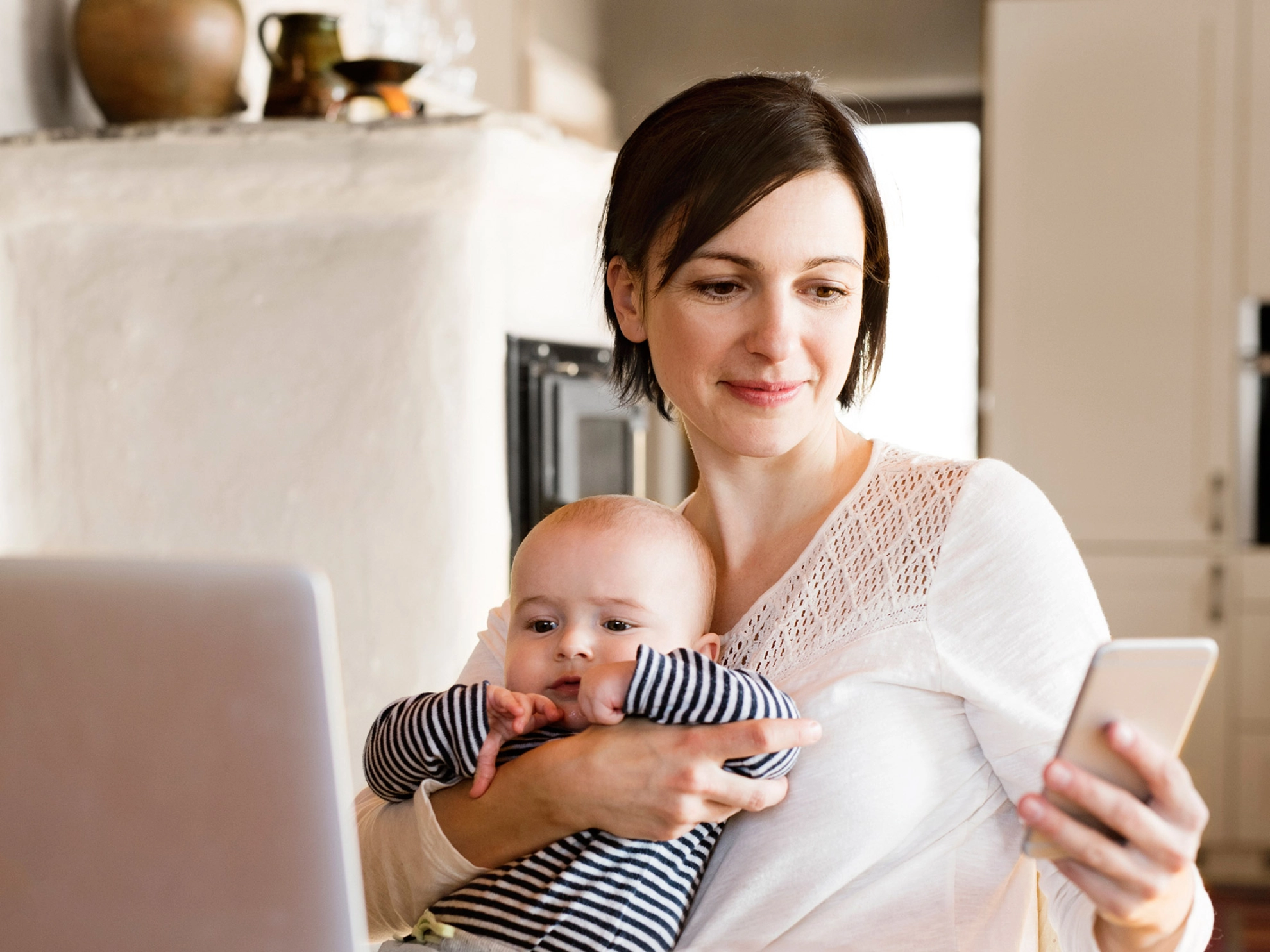 Eine junge Frau sitzt am Küchentisch und telefoniert. Vor ihr steht eine Tasse und ein geschlossenes Notebook.
